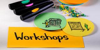 Foto mit Moderationsmaterialien: Im Vordergrund eine rechteckige Metaplankarte mit dem Wort "Workshops", rechts darüber zwei runde Metaplankarten mit gezeichneten Workshopszenen, darüber Moderationsstifte, Pinnadeln und ein Stapel runde Metaplankarten
