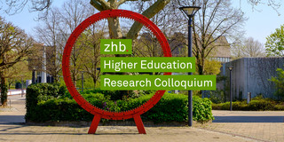Photo of the red gear wheel on Martin-Schmeißer-Platz at TU Dortmund University. The lettering zhb Higher Education Research Colloquium protrudes from the gear wheel from left to right