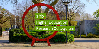 zhb Higher Education Research Colloquium Photo of the red gear wheel on Martin-Schmeißer-Platz at TU Dortmund University. The lettering zhb Higher Education Research Colloquium protrudes from the gear wheel from left to right