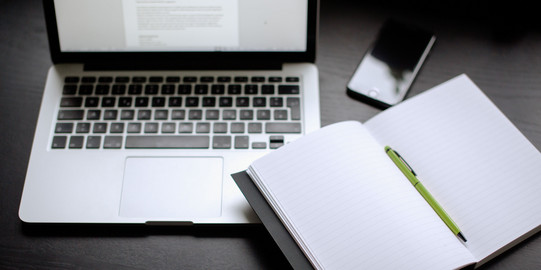 Laptop, Smartphone und Notizbuch mit Stift auf einem Tisch liegend.