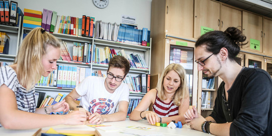 Vier Personen sitzen an einem Tisch und beschäftigen sich mit Bausteinen, die auf dem Tisch liegen. Im Hintergund steht ein Regal voller Bücher und Ordner und ein Schrank.