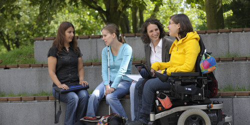 Vier Studierende, darunter Studierende mit Beeinträchtigung, sitzen gemeinsam auf dem Campus.