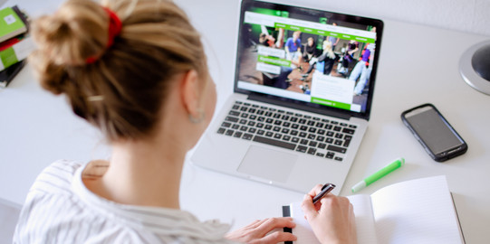 Am Schreibtisch sitzende Studentin arbeitet am Laptop, daneben liegen Bücher und Stifte.