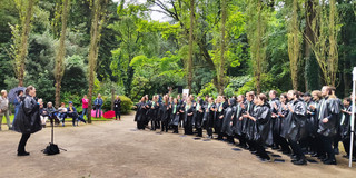 Auftritt des Unichors am Pappelrondell im Rombergpark. Links im Bild die Chorleiterin, rechts die Chorgruppe. Alle in schwarzen Regencapes und grünen TU-Halstüchern bzw. Krawatten.