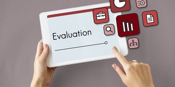 Abgebildet ist ein Tablet, auf dem 'Evaluation' und damit verbundenen Symbolen zu sehen ist, sowie zwei Hände. 