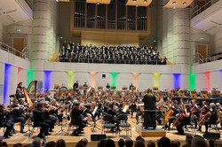 Heinke Kirzinger dirigiert die Zugabe. Vorne sitzt das Orchester. Auf der Empore steht der Chor.