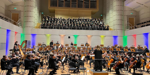 Heinke Kirzinger dirigiert die Zugabe. Vorne sitzt das Orchester. Auf der Empore steht der Chor.