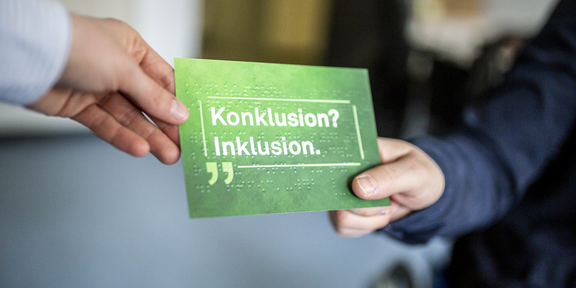Eine grüne Postkarte mit der Aufschrift „Konklusion? Inklusion.“. Der Schriftzug ist zusätzlich in Brailleschrift in die Postkarte geprägt. An der oberen linken Ecke der Postkarte eine Hand, die die Postkarte an eine Person übergibt, die die rechte Ecke hält. Im Hintergrund das Rad eines Rollstuhls.