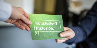 Eine grüne Postkarte mit der Aufschrift „Konklusion? Inklusion.“. Der Schriftzug ist zusätzlich in Brailleschrift in die Postkarte geprägt. An der oberen linken Ecke der Postkarte eine Hand, die die Postkarte an eine Person übergibt, die die rechte Ecke hält. Im Hintergrund das Rad eines Rollstuhls.