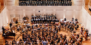 Auftritt des Unichors im Dortmunder Konzerthaus: Die Sänger*innen stehen auf der Chorempore und im hinteren Teil der Bühne. Auf der Bühne ist ein Orchester. Vorne in der Mitte steht die Dirigentin.