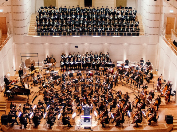 Auftritt des Unichors im Dortmunder Konzerthaus: Die Sänger*innen stehen auf der Chorempore und im hinteren Teil der Bühne. Auf der Bühne ist ein Orchester. Vorne in der Mitte steht die Dirigentin.