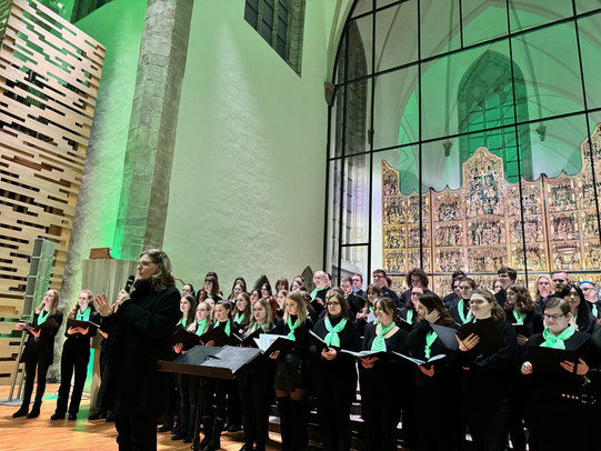 Der Unichor steht vor dem Altar der Sankt Petri Kirche, vorne steht Heinke Kirzinger mit Mikrofon in der Hand.