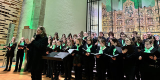 Der Unichor steht vor dem Altar der Sankt Petri Kirche, vorne steht Heinke Kirzinger mit Mikrofon in der Hand.