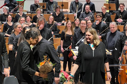 Gratulation an Dirigent und Dirigentin mit Blumensträußen. Im Hintergrund steht das Orchester.
